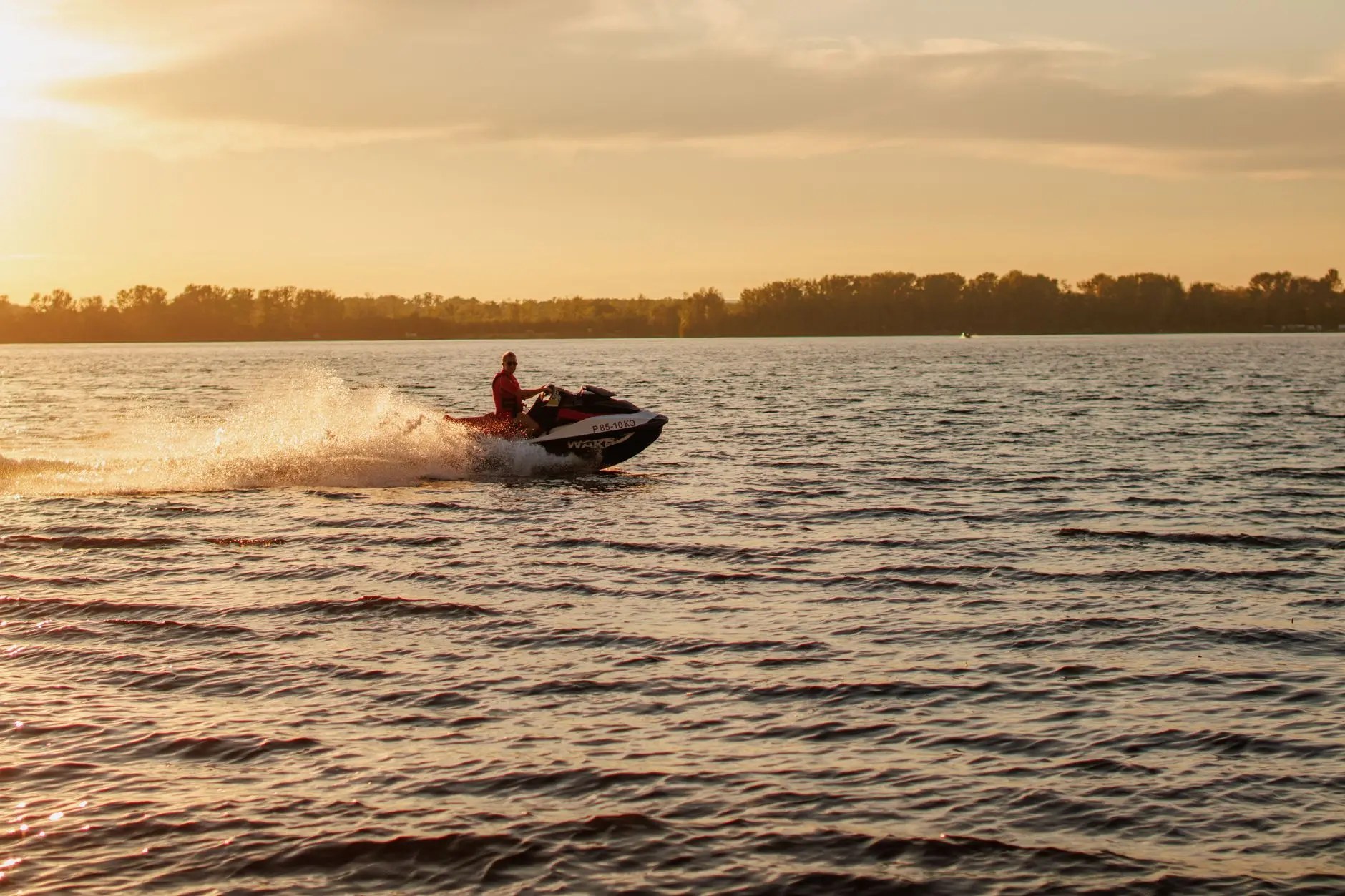 A jet ski speeds across a lake at sunset, with water splashing behind.
