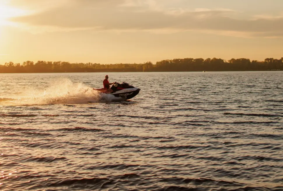 A jet ski speeds across a lake at sunset, with water splashing behind.