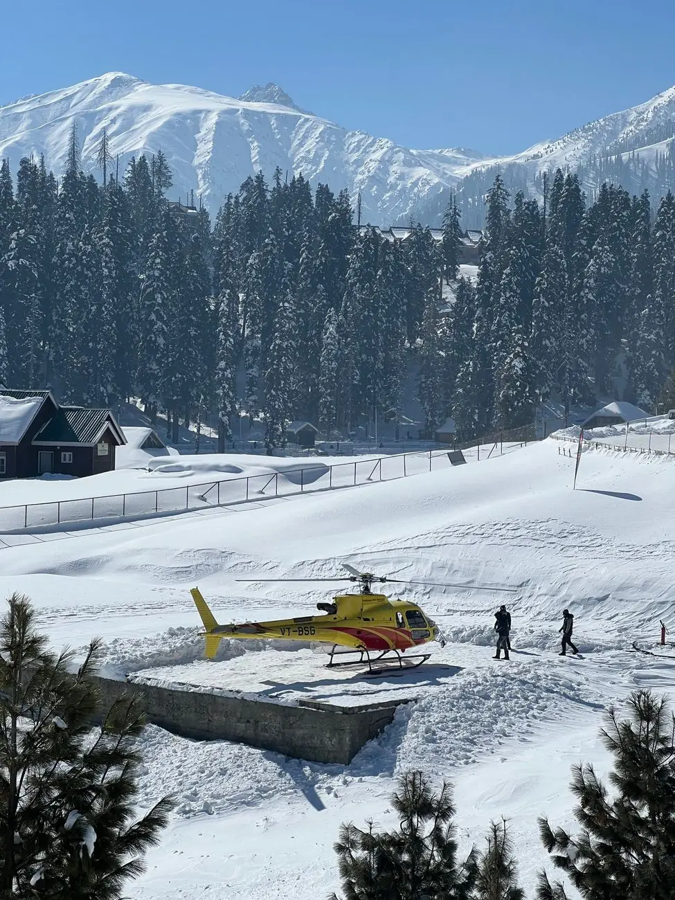 Yellow helicopter parked on snowy Gulmarg backdrop with majestic mountains and pine trees.